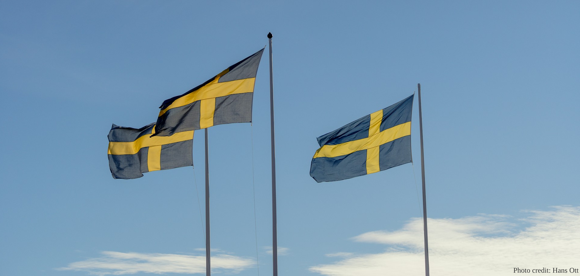 Three Swedish national flags, featuring a yellow Nordic cross on a bright blue field, fly from tall white flagpoles against a clear, pale blue sky. The photo is taken from a low angle, looking upward, with a few wispy clouds visible at the bottom right. A watermark in the bottom right corner credits the photo to Hans Ott.