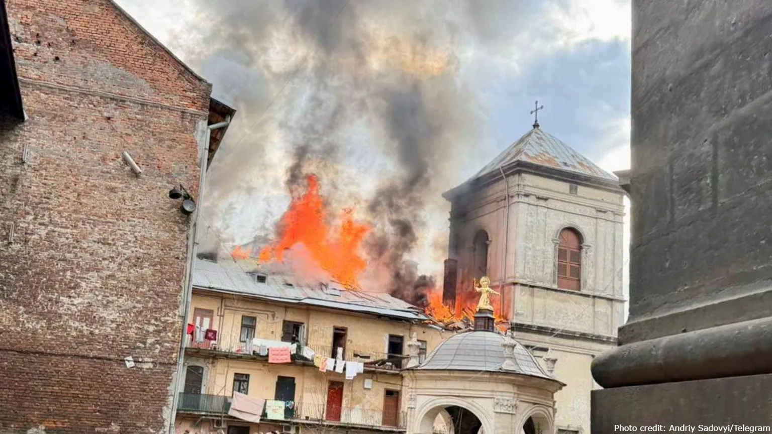A large fire with intense orange flames and thick black and grey smoke billows from the roof of a multi-story historic building within the Bernardine Monastery complex in Lviv. The 17th-century monastery tower and a smaller dome topped with a golden statue are visible in the background and foreground, respectively. The scene, captured on March 24, 2026, shows the aftermath of a major Russian Shahed drone strike on the city's historic center, which is a UNESCO World Heritage site. Photo credit: Andriy Sadovyi/Telegram.