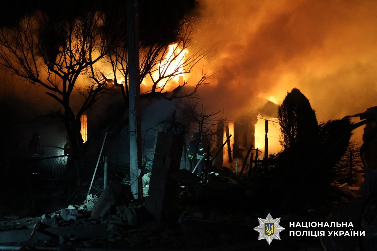 A house is engulfed in intense orange flames and thick smoke at night, with the silhouettes of trees and a utility pole in the foreground. The National Police of Ukraine logo is visible in the bottom right corner.