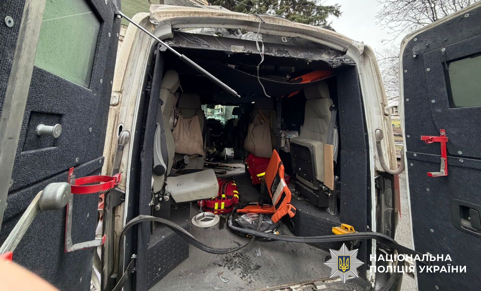 The damaged interior of a vehicle is shown through its open rear doors. The roof and upper frame of the doorway are mangled and charred, while red medical bags and orange rescue equipment are visible inside near the seats. The National Police of Ukraine logo is in the bottom right corner.