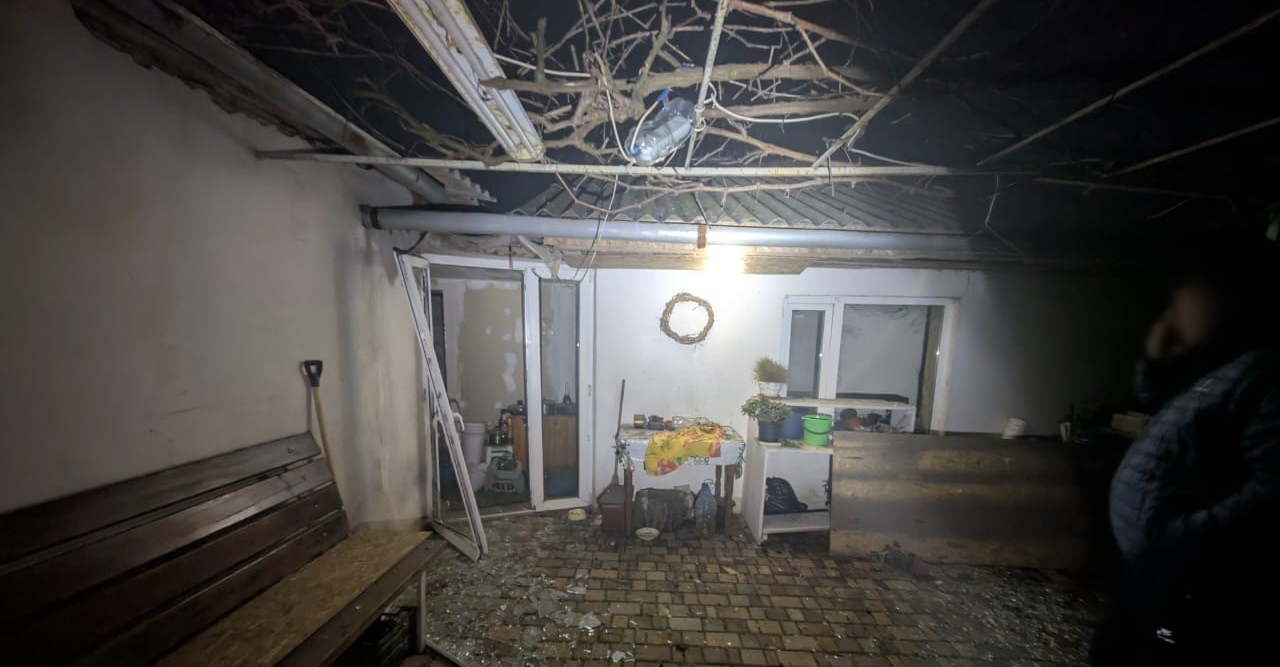The courtyard of a private residence at night is blanketed in shattered glass and debris following a Russian strike in the Zaporizhzhia region. A wooden bench stands on the left, and a table with scattered household items is visible in the center. The National Police of Ukraine logo is in the bottom-right corner, documenting the aftermath of the heavy shelling