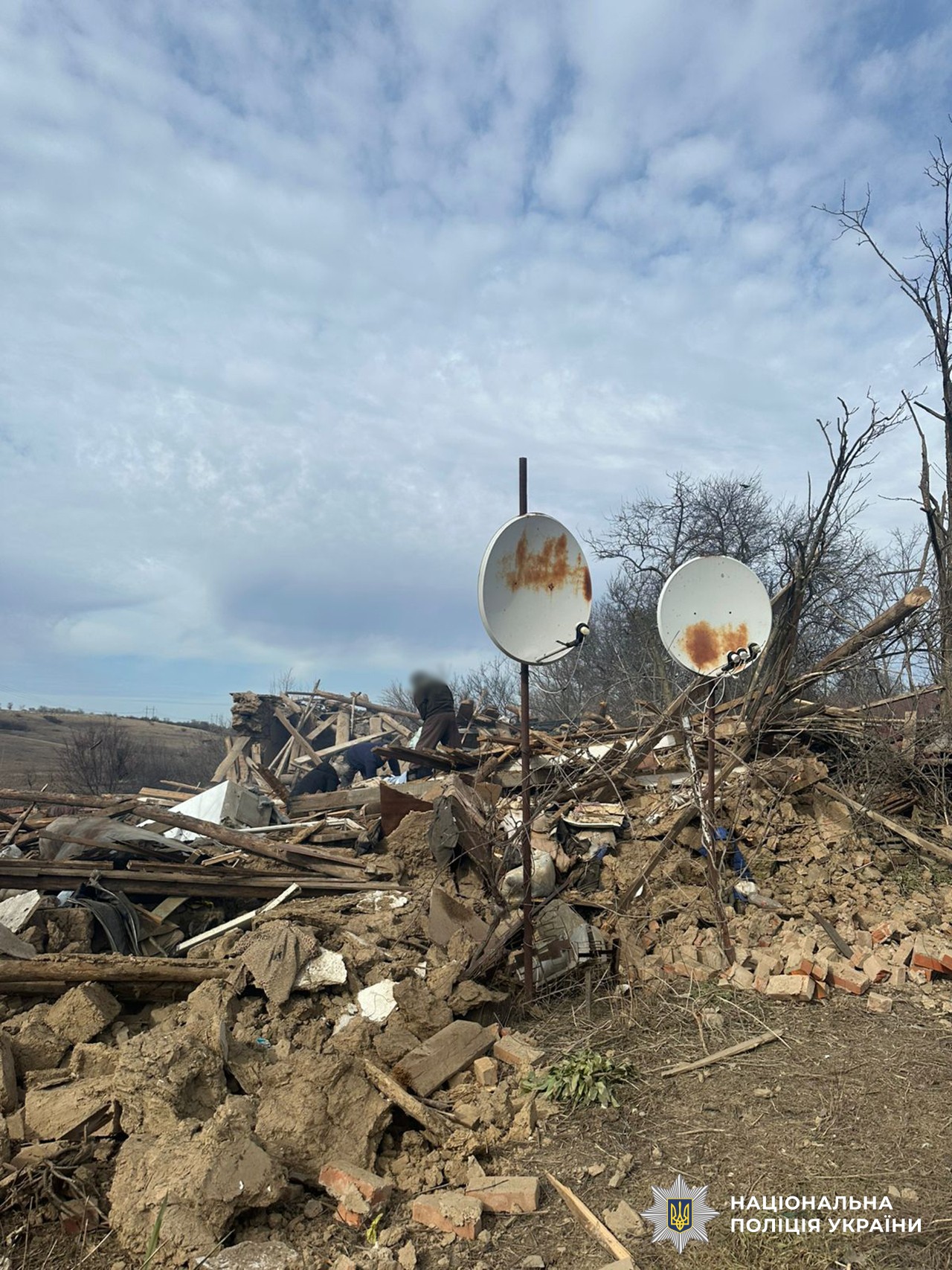 A massive pile of rubble from a completely destroyed building dominates the landscape under a cloudy sky. Two white satellite dishes, both showing significant rusty patches, are mounted on metal poles in the foreground. In the middle ground, several people with blurred faces are seen working or searching through the debris of wood, bricks, and concrete. The National Police of Ukraine logo is visible in the bottom-right corner.