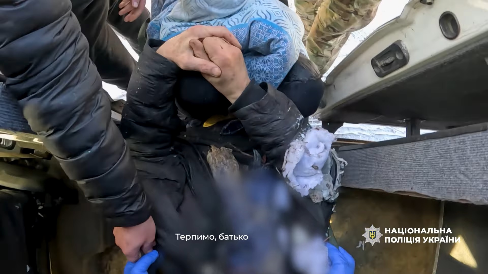 A close-up, high-angle view shows first responders in blue gloves and tactical gear carefully assisting an injured person wrapped in a blue patterned blanket. The National Police of Ukraine logo is in the bottom right, and a subtitle in Ukrainian reads, "Hang in there, father"