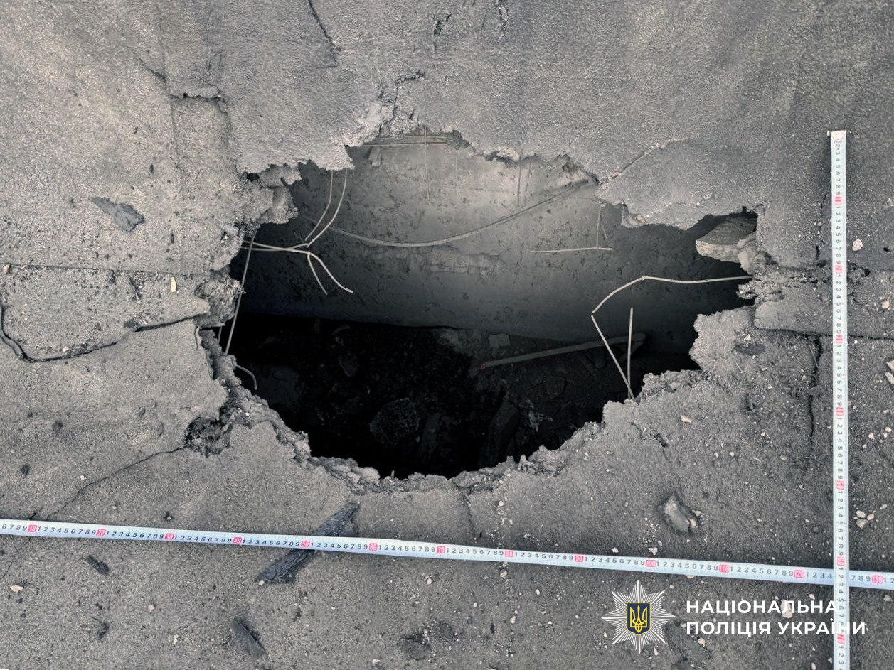 A top-down close-up photo shows a large, jagged hole in a grey concrete surface,  caused by an explosion. Inside the hole, twisted metal reinforcement bars (rebar) and dark debris are visible. Two white measuring tapes with red and black markings are laid out horizontally and vertically across the crater to document its size. In the bottom right corner, the official logo of the National Police of Ukraine (Національна поліція України) is displayed.