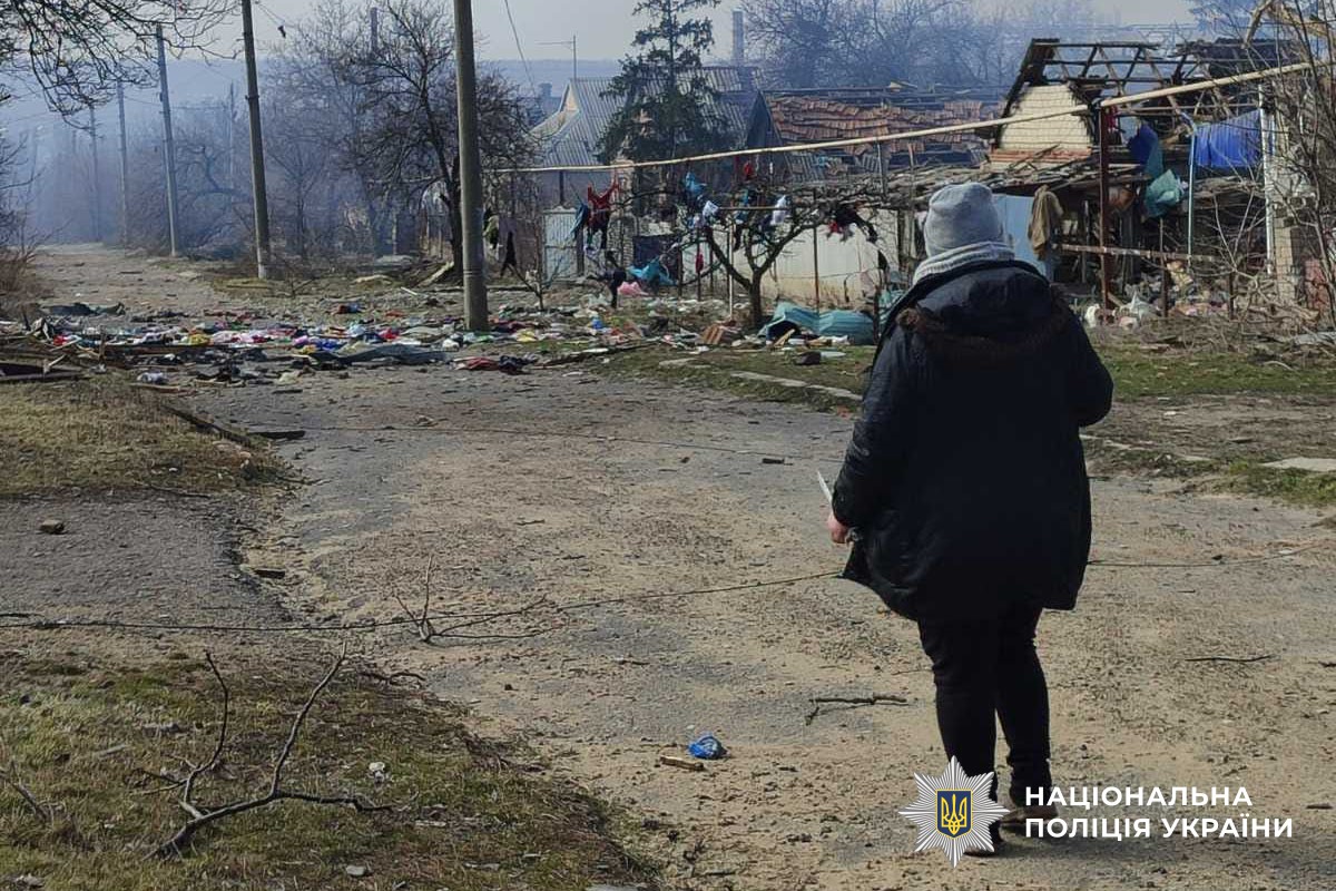 A woman in a dark coat and winter hat stands on a dirt road, looking toward a village street cluttered with debris and damaged houses. The National Police of Ukraine logo is visible in the bottom right corner.