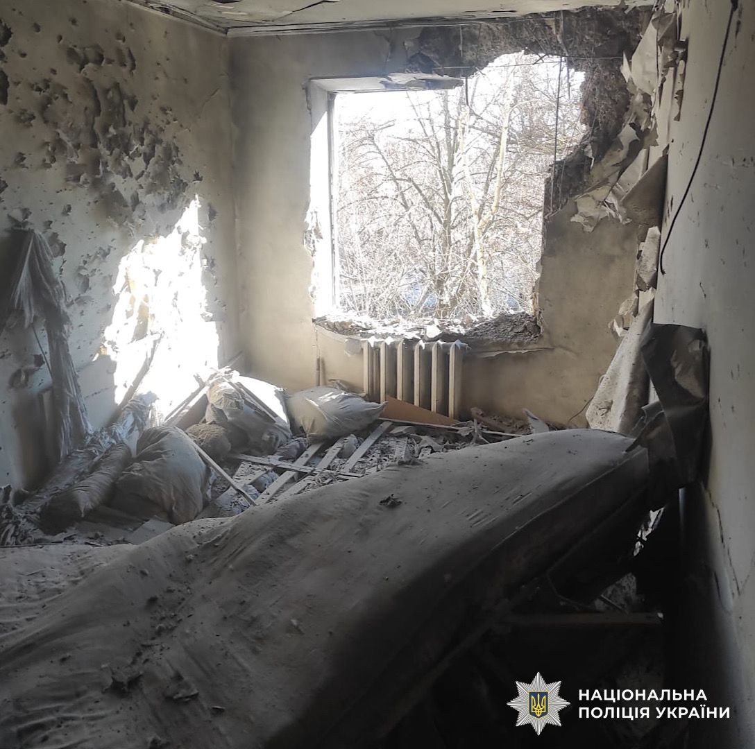 The interior of a room is shown heavily damaged with a large hole in the wall, shrapnel-pockmarked walls, and furniture covered in thick dust and debris. The National Police of Ukraine logo is in the bottom right corner.