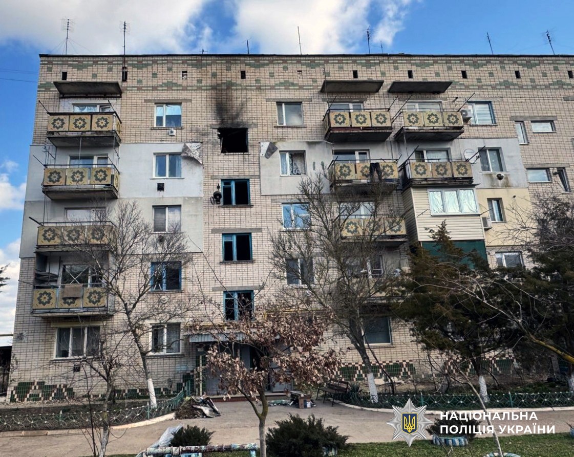 A five-story brick apartment building shows damage from a strike, with a charred, blackened window on the fourth floor and several other shattered windows. The building features balconies with a yellow and green floral pattern, and the National Police of Ukraine logo is visible in the bottom right corner.