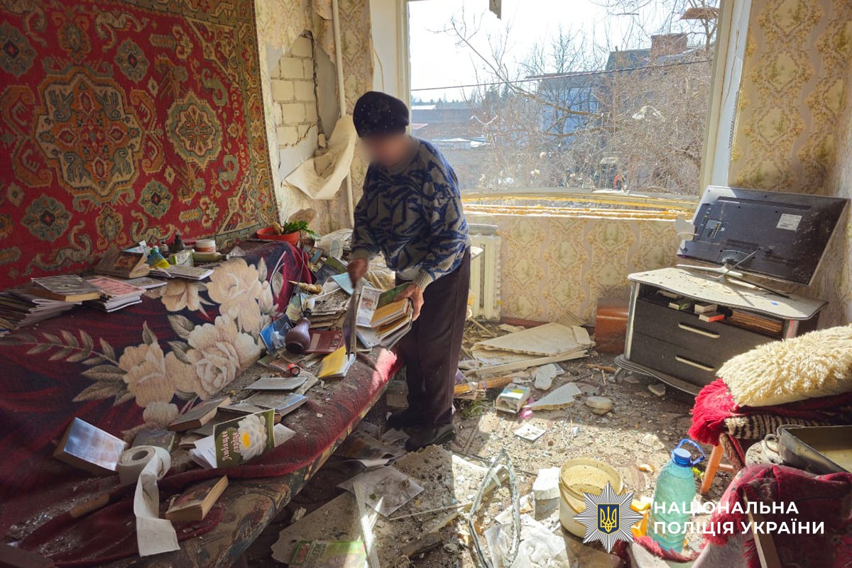 An elderly person stands in a severely damaged room filled with debris, holding books near a sofa covered in dust and fallen plaster. The window is completely blown out, and the National Police of Ukraine logo is visible in the bottom right corner.