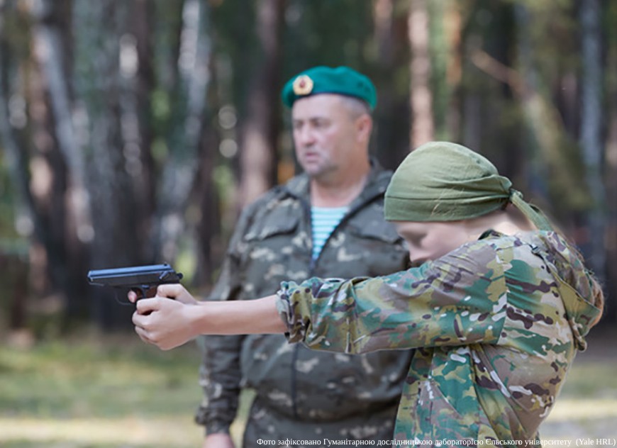 A close-up shot of a young person with a blurred face, wearing a camouflage jacket and an olive-green headscarf, aiming a black handgun with both hands. Standing slightly behind and supervising is an adult man in a camouflage jacket and a green military beret. The training takes place in a blurred forest environment. A watermark at the bottom right attributes the photo to the Yale University Humanitarian Research Lab (Yale HRL).