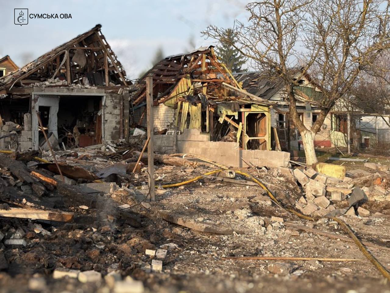 Severely damaged residential houses in Sumy Oblast stand in ruins with collapsed roofs and exposed wooden frames following an attack. Bricks, splintered wood, and household debris cover the ground in the foreground, while the Sumy Regional Military Administration (OVA) logo is visible in the upper-left corner.