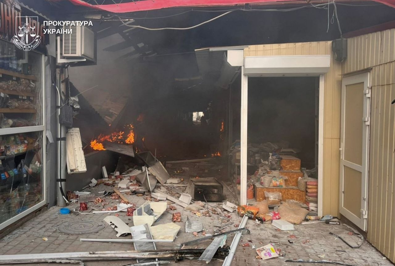 The entrance to a small marketplace or shop is destroyed, with debris and scattered goods littering the pavement in the foreground. Small fires burn within the dark, smoke-filled interior of the structure, and the logo for the Prosecutor General's Office of Ukraine is visible in the top-left corner.