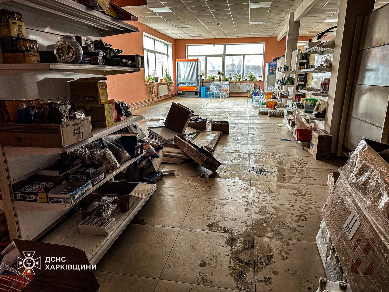 The interior of a grocery store in Kharkiv Oblast shows water-damaged floors and scattered cardboard boxes in the main aisle. Shelves remain stocked with snacks and household goods, while large windows at the back let in natural light, and the State Emergency Service of Ukraine (DSNS) logo is visible in the bottom-left corner.