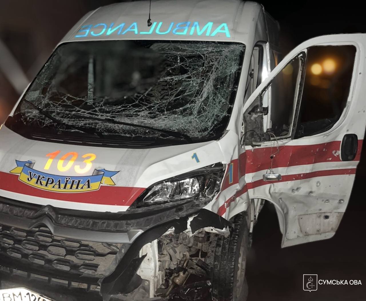 A Ukrainian ambulance in Sumy Oblast shows heavy damage from an attack at night, with its windshield completely shattered and the front driver's side crumpled. Shrapnel holes are visible along the side door and body of the white vehicle, which features red stripes and the "103" emergency number, while the Sumy Regional Military Administration (OVA) logo appears in the bottom-right corner.