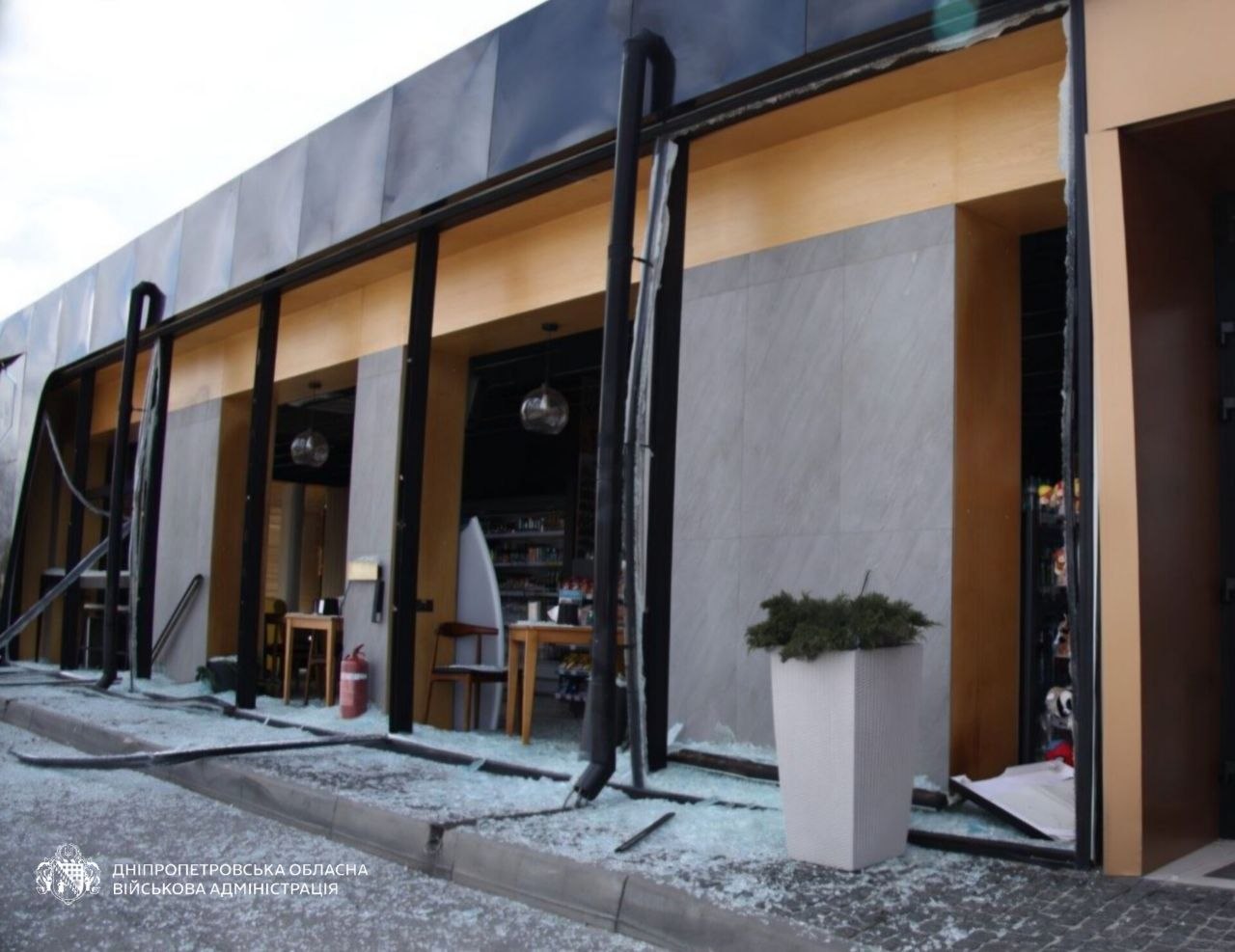 The glass storefront of a modern commercial building in Dnipropetrovsk Oblast is completely shattered, with shards of glass covering the sidewalk and interior floor. Bent black gutters hang from the facade, exposing the tables and shelving inside the shop, while the Dnipropetrovsk Regional Military Administration logo is visible in the lower-left corner.