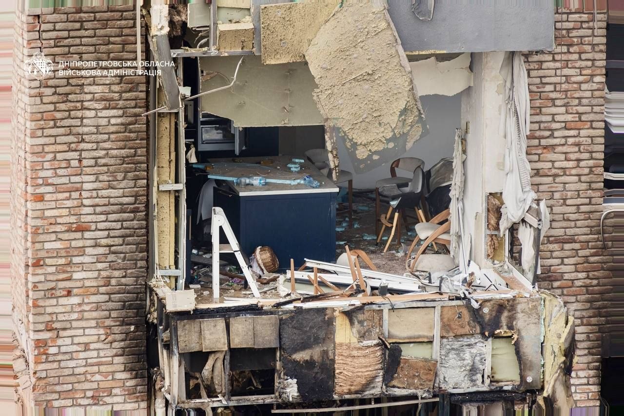 The exterior of a brick apartment building in Dnipropetrovsk Oblast shows a large, gaping hole where a wall and window have been blown out, exposing a modern kitchen and dining area inside. Debris from the ceiling and facade hangs precariously over a dark blue kitchen island and chairs, with the Dnipropetrovsk Regional Military Administration logo visible in the upper-left corner.