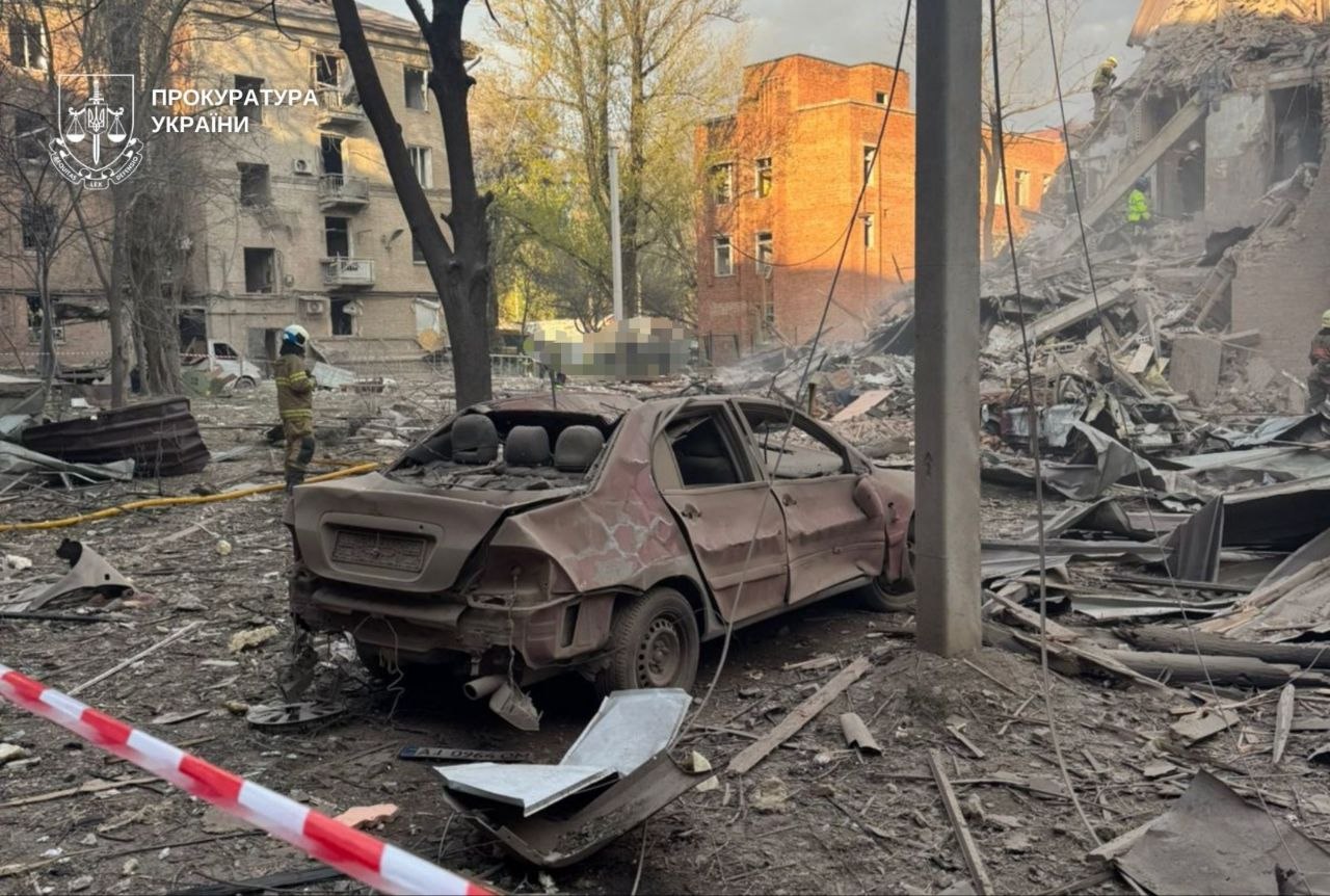 A damaged, dust-covered car sits in the center of a debris-strewn street following an explosion. In the background, rescue workers in uniform navigate a massive pile of rubble where a building has completely collapsed. To the left, an apartment building shows blown-out windows and structural damage. Red and white police tape cordons off the area in the foreground, and the logo of the Prosecutor's Office of Ukraine is visible in the top-left corner.