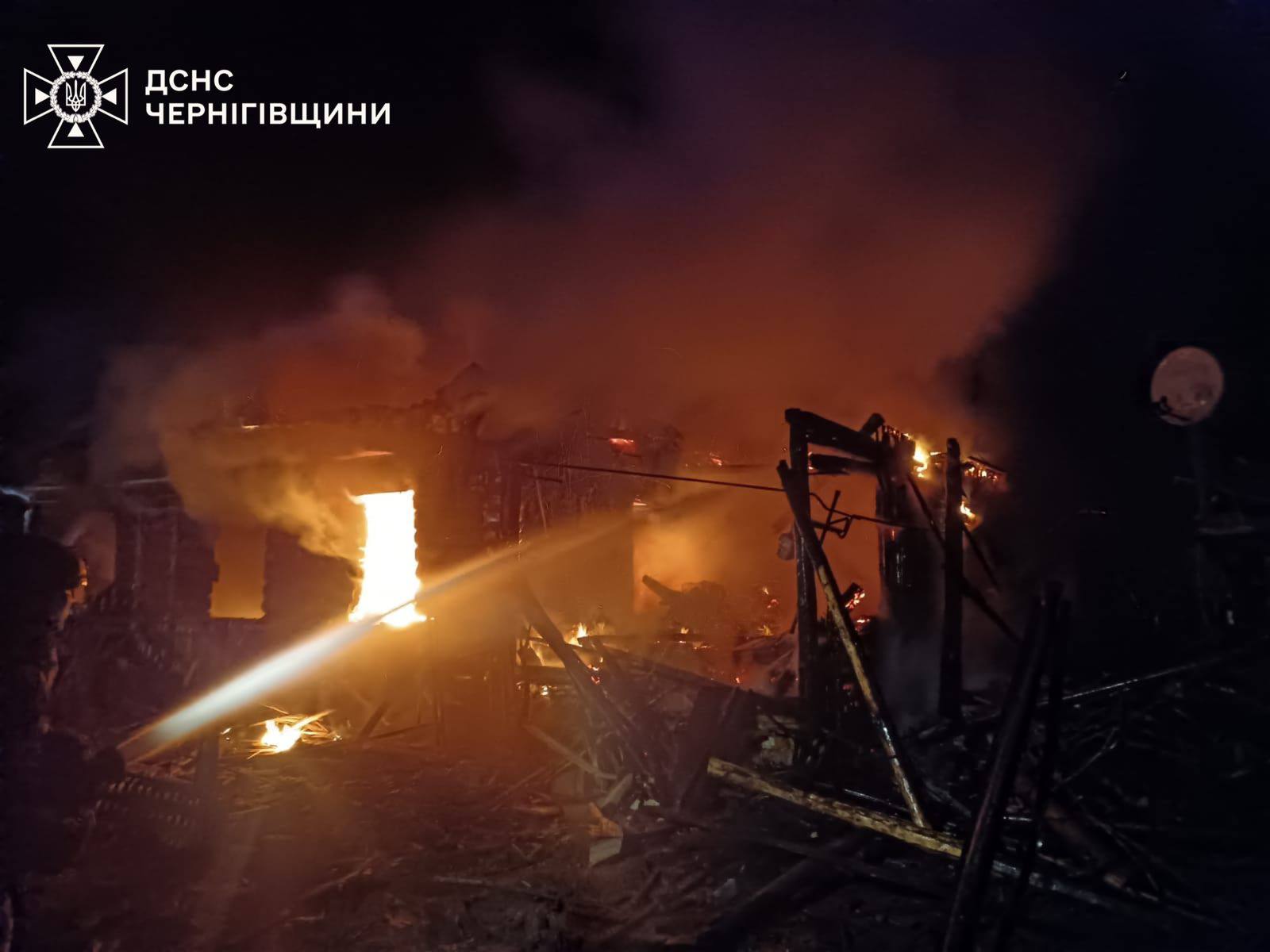 A nighttime scene in Chernihiv Oblast shows firefighters battling a large structural fire that has reduced a building to charred wooden skeletons. A powerful jet of water from a fire hose cuts through the darkness, aimed at the glowing orange flames and thick smoke rising from the debris. The State Emergency Service of Ukraine (DSNS) logo for the Chernihiv region is visible in the top-left corner.