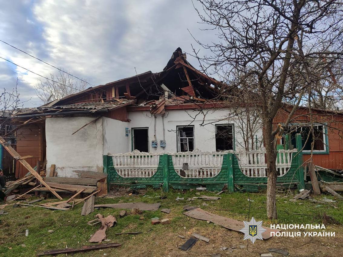 A small, one-story residential house shows significant damage following an attack, with a large portion of the slate roof collapsed and the wooden supporting structure exposed. The front windows are blown out, and debris is scattered across the yard in front of a green and white decorative concrete fence. The National Police of Ukraine logo is visible in the bottom-right corner.