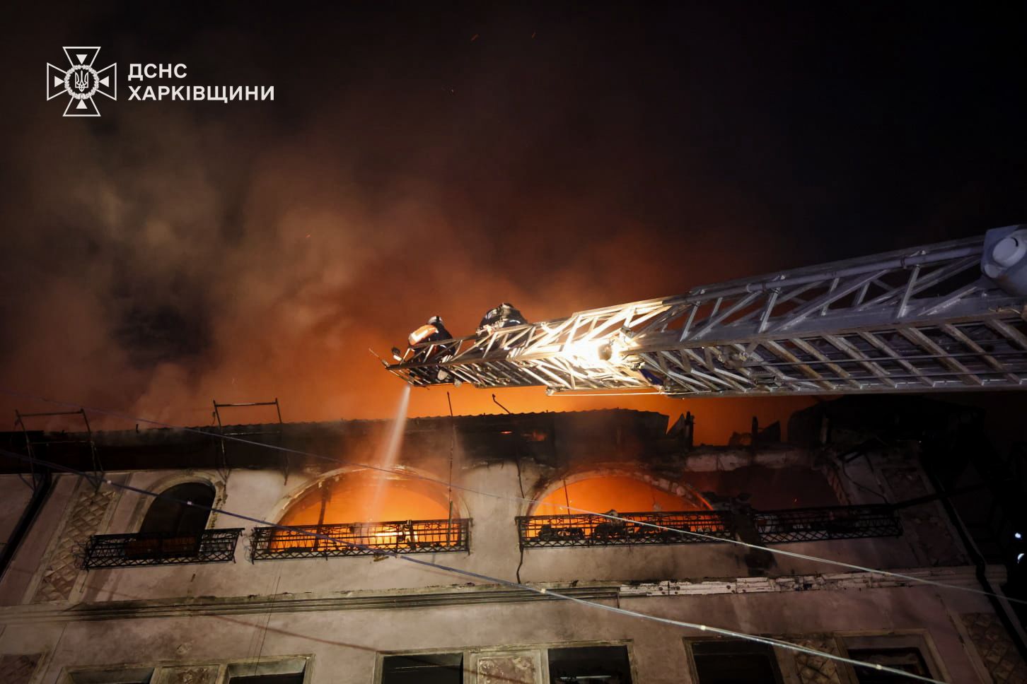 Firefighters in Kharkiv Oblast use an aerial ladder to direct a water stream into the arched windows of a burning building at night. Intense orange flames glow from within the structure, illuminating smoke rising into the dark sky, with the State Emergency Service of Ukraine (DSNS) logo in the upper-left corner.