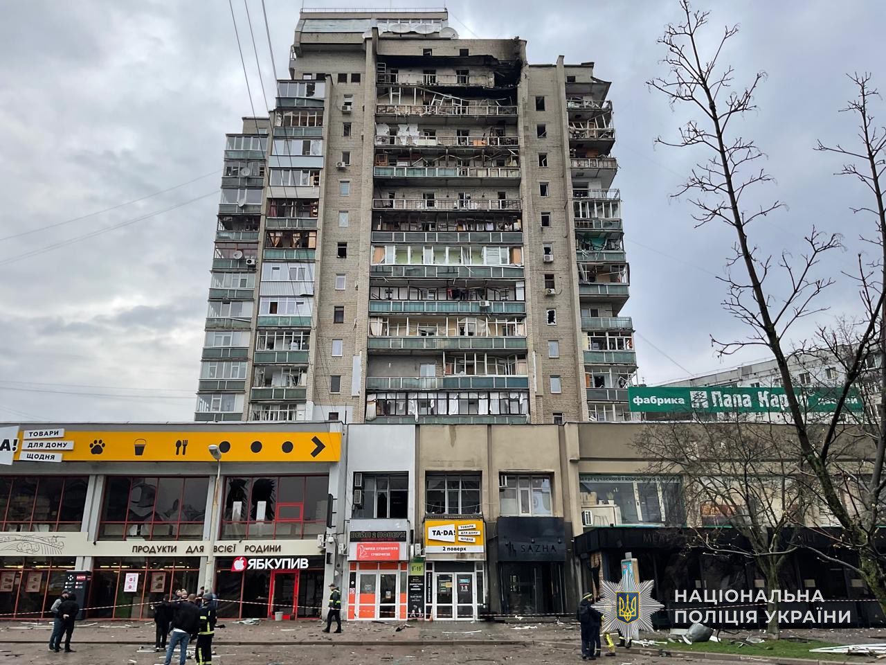 A tall residential apartment building shows extensive damage to several upper-floor balconies and windows, with scorched sections near the roof. Below the residential units, ground-level storefronts feature shattered windows and cordoned-off areas where police and emergency personnel are gathered, with the National Police of Ukraine logo in the bottom right corner.