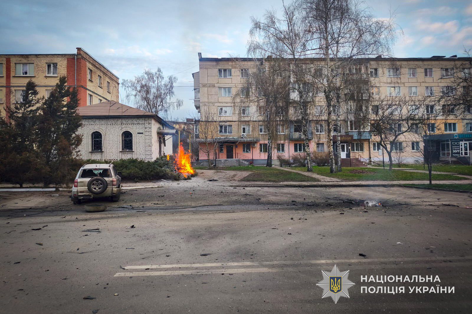 Alt-text Generator Custom Gem Alt-text Generator said A street scene reveals the aftermath of an attack, with debris scattered across the asphalt and a small fire burning in a bush near a residential building. A damaged silver SUV with a shattered rear window sits parked nearby, while apartment blocks with several broken windows stand in the background, accompanied by the National Police of Ukraine logo.
