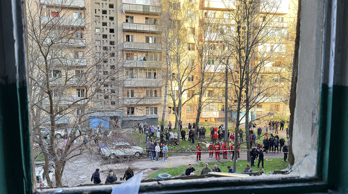 Viewed through a shattered window frame with jagged glass, a large group of civilians, police officers, and emergency responders in red uniforms gather in the courtyard of a multi-story apartment building. The area is cordoned off with red and white tape, and the National Police of Ukraine logo is visible in the lower-right corner.