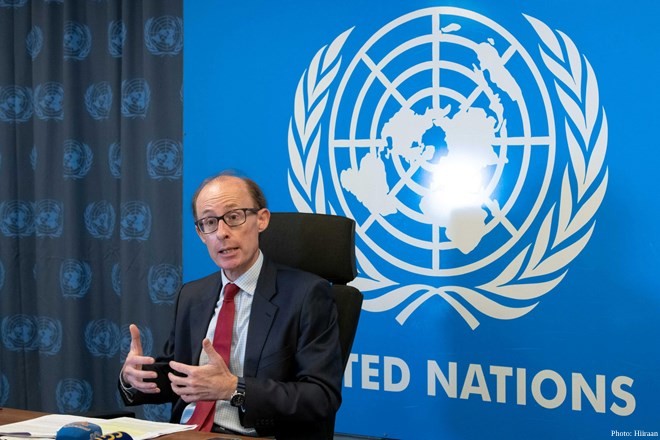 Ben Saul, the UN Special Rapporteur on human rights and counter-terrorism, sits at a desk and gestures with his hands while speaking. He is wearing a dark suit, white shirt, and red tie, positioned in front of a large blue backdrop featuring the United Nations logo and text. A grey curtain with a repeating UN emblem is visible to the left.