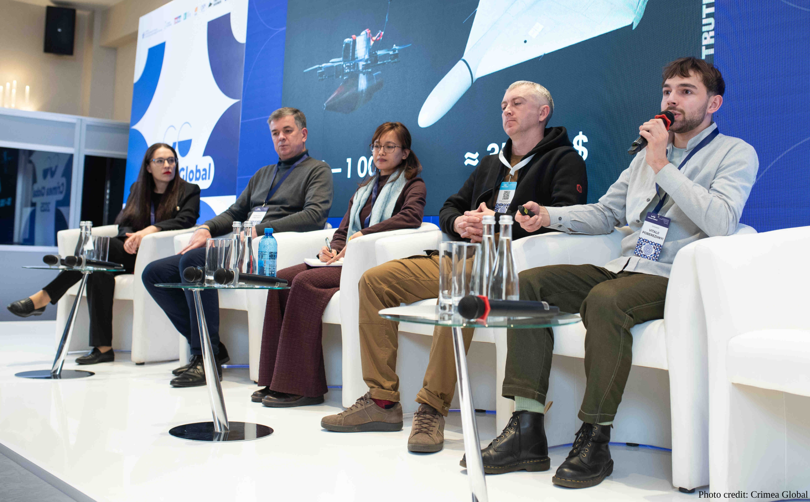 Five panelists sit in white armchairs on a stage during a "Crimea Global" conference, with one man on the far right speaking into a microphone. A large digital screen behind them displays graphics of drones and text, while small glass tables with water bottles and microphones are positioned in front of the participants.