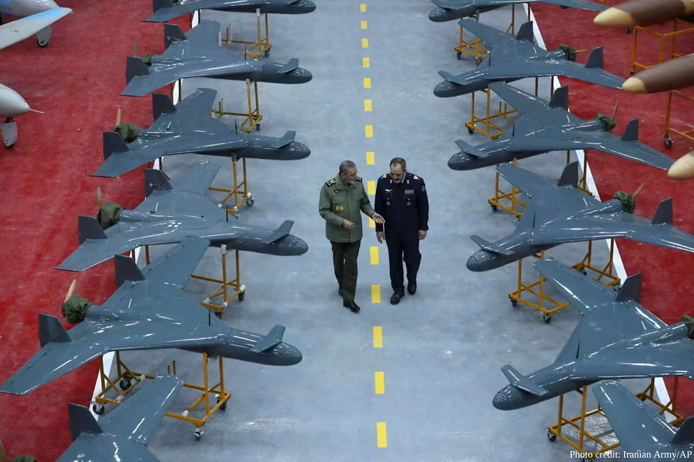 Two high-ranking military officials in uniform walk down a central aisle between rows of grey delta-wing suicide drones, Shahed models, mounted on yellow transport stands. The facility has a light blue floor with a dashed yellow line, and the photo is credited to the Iranian Army/AP in the bottom-right corner.