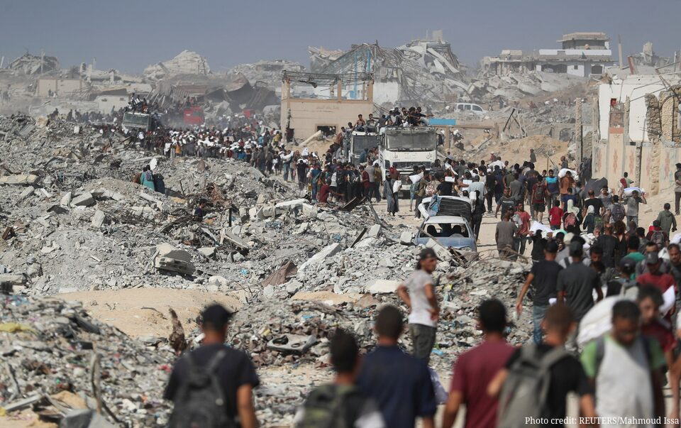 A large crowd of Palestinians, many carrying white bags of aid supplies, walk through a landscape of total devastation in Beit Lahia, northern Gaza. Destroyed buildings and massive piles of grey rubble line the dusty road, where aid trucks carrying people on their roofs are also visible. The photo, taken on August 2, 2025, is credited to REUTERS/Mahmoud Issa in the bottom-right corner.