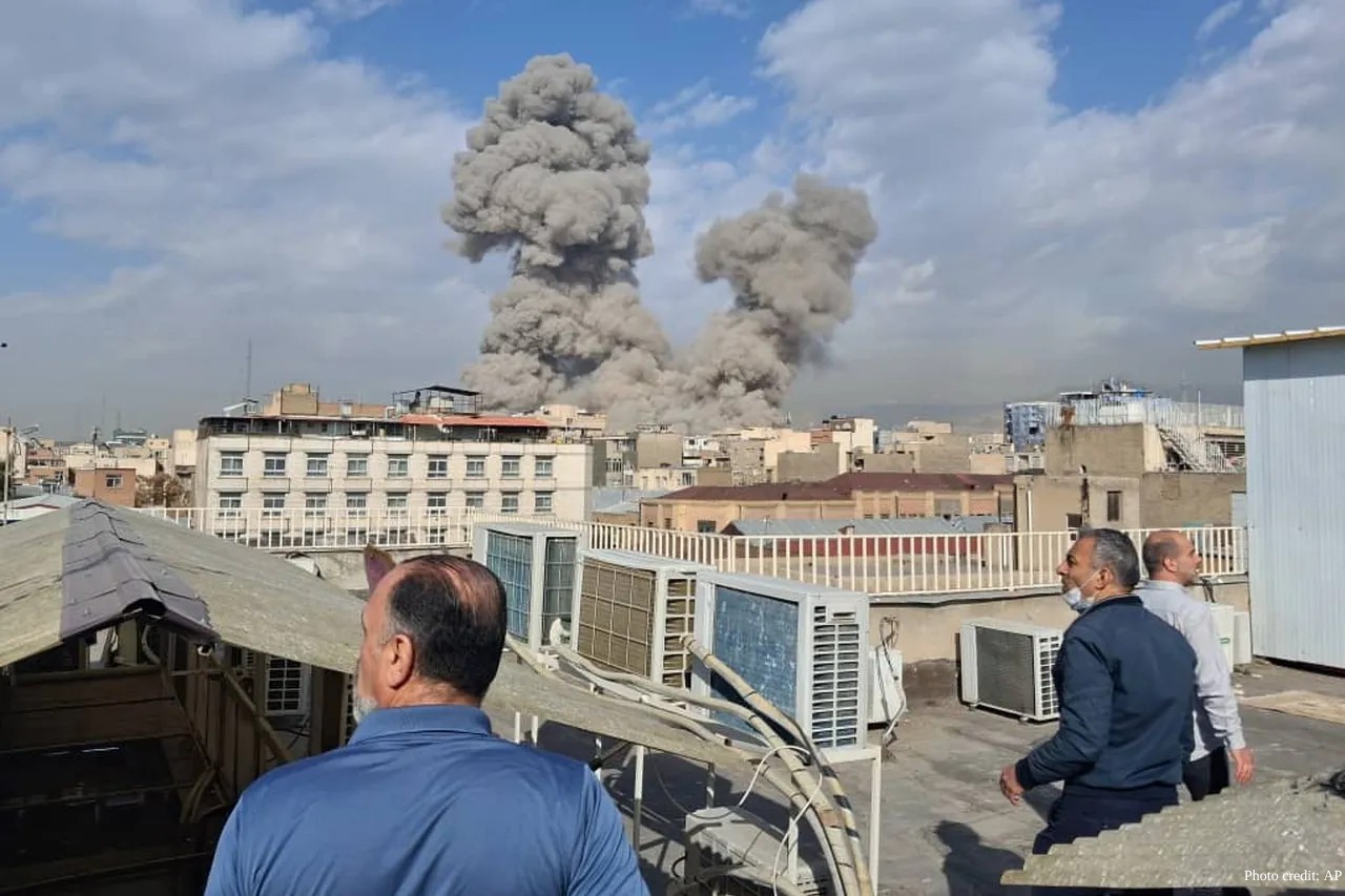 From a rooftop in Tehran, several men watch as two massive, mushroom-shaped plumes of thick grey smoke billow into the sky following an explosion on February 28, 2026. The foreground shows air conditioning units and building structures, while the background reveals a dense cityscape under a bright, partly cloudy sky. The photo is credited to AP in the bottom-right corner.