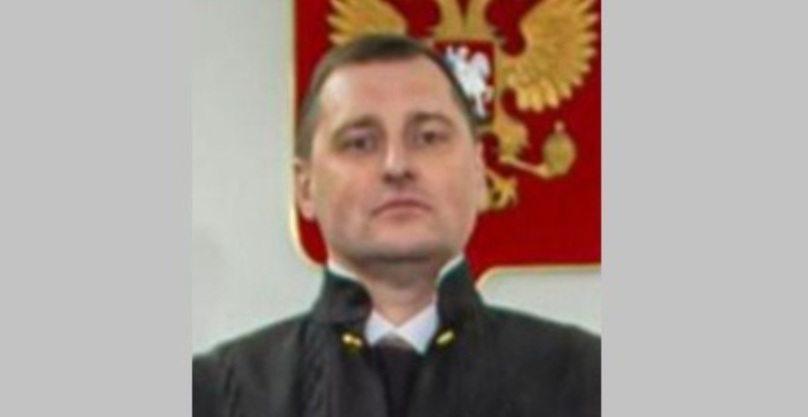 A portrait of a man with short brown hair, wearing a black judicial robe with a high collar over a white shirt and dark tie. He is positioned in front of a wall featuring the coat of arms of the Russian Federation — a golden double-headed eagle on a red shield.