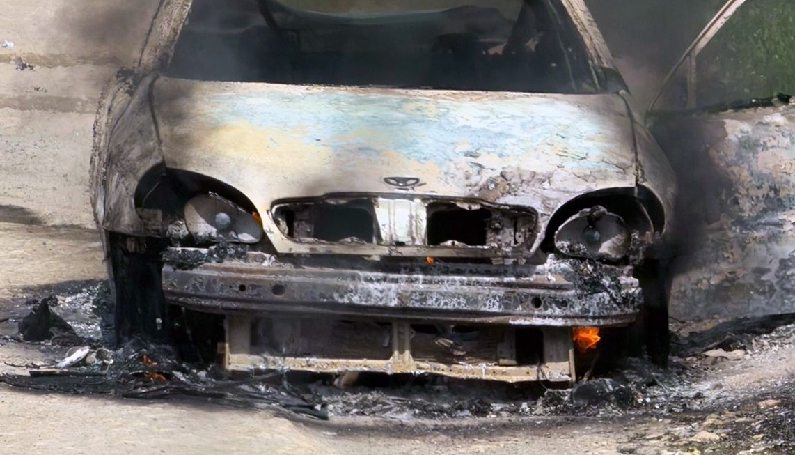 A close-up front view of a Daewoo Lanos sedan that has been entirely destroyed by fire. The car’s body is heavily charred and blackened, with all its internal components—engine bay, dashboard, and seats—missing or melted. Thick, acrid smoke continues to billow from the vehicle as small flames still flicker on the ground beneath the engine and chassis. The National Police of Ukraine logo is visible in the bottom-right corner.