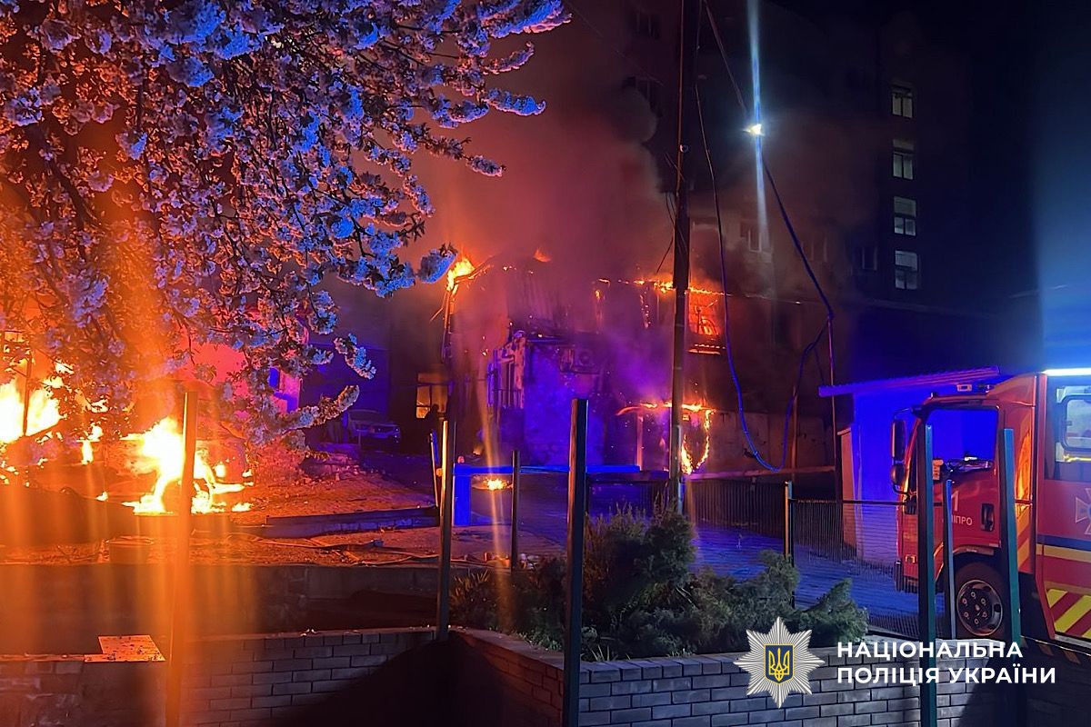 A night scene in Dnipropetrovsk Oblast shows a building engulfed in intense orange flames and thick smoke following an attack. A flowering tree in the foreground is illuminated by the fire, while a red fire engine is parked on the right, and the National Police of Ukraine logo is in the bottom-right corner.