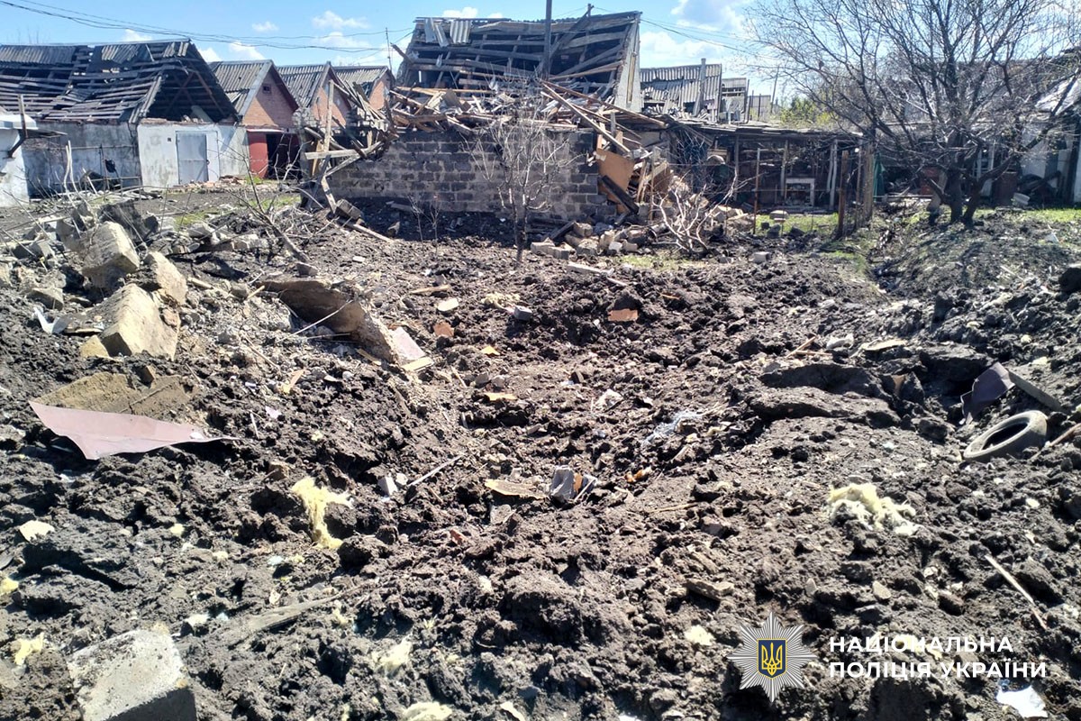 A large impact crater in dark, churned earth dominates the foreground of a residential yard. In the background, several small outbuildings and houses show significant structural damage, with collapsed roofs and exposed wooden beams. Debris, including bricks and insulation, is scattered across the site. The National Police of Ukraine logo is visible in the bottom-right corner.