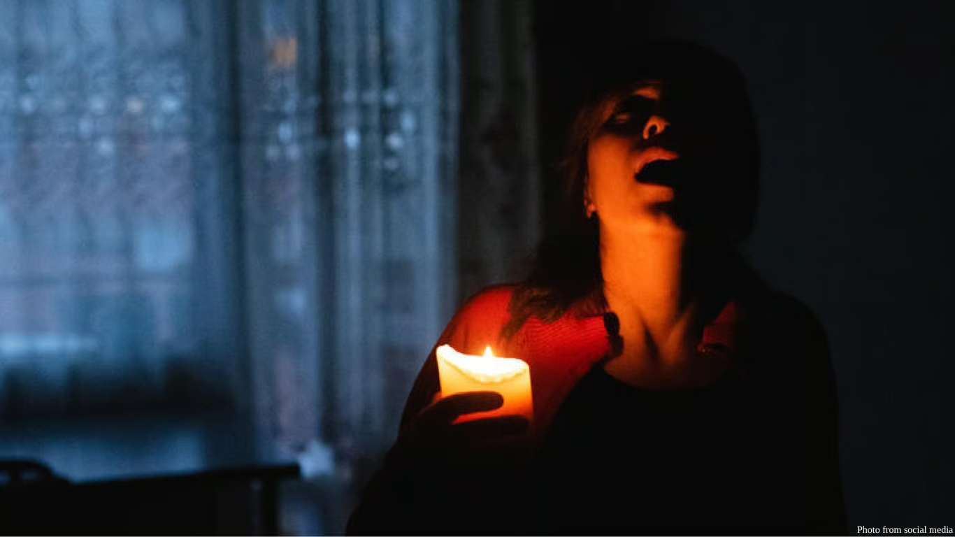 A cinematic, low-light shot of a woman in a dark room, her face partially illuminated by the warm glow of a single lit candle she is holding. She has an anguished expression, with her mouth open as if crying out or grieving. The background shows a window with sheer curtains in dim, blueish light. A small "Photo from social media" credit is in the bottom-right corner.