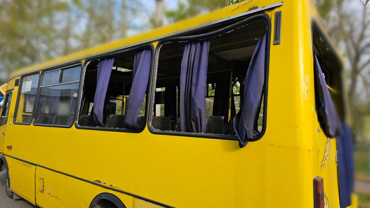 A bright yellow bus is shown with its side windows completely shattered. Dark blue curtains hang limply from the empty window frames, some torn and flapping. The upper metal frame of the windows is slightly warped, and the background consists of blurred green trees.