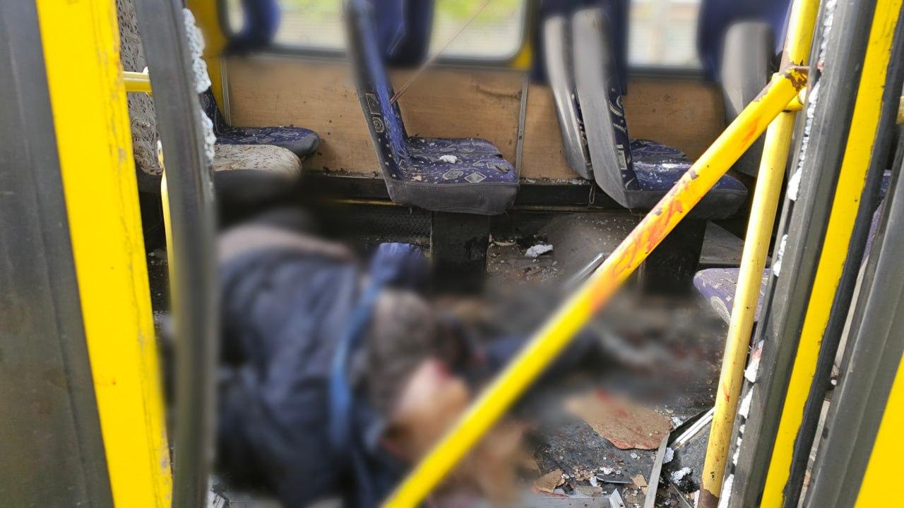 A view from the doorway of a yellow bus shows the interior covered in debris and shattered glass. In the center of the aisle, a human figure lies on the floor; the body and immediate surrounding area are heavily blurred. The bus seats are patterned blue, and some appear to have shrapnel damage or debris on them. Yellow handrails frame the foreground of the shot.