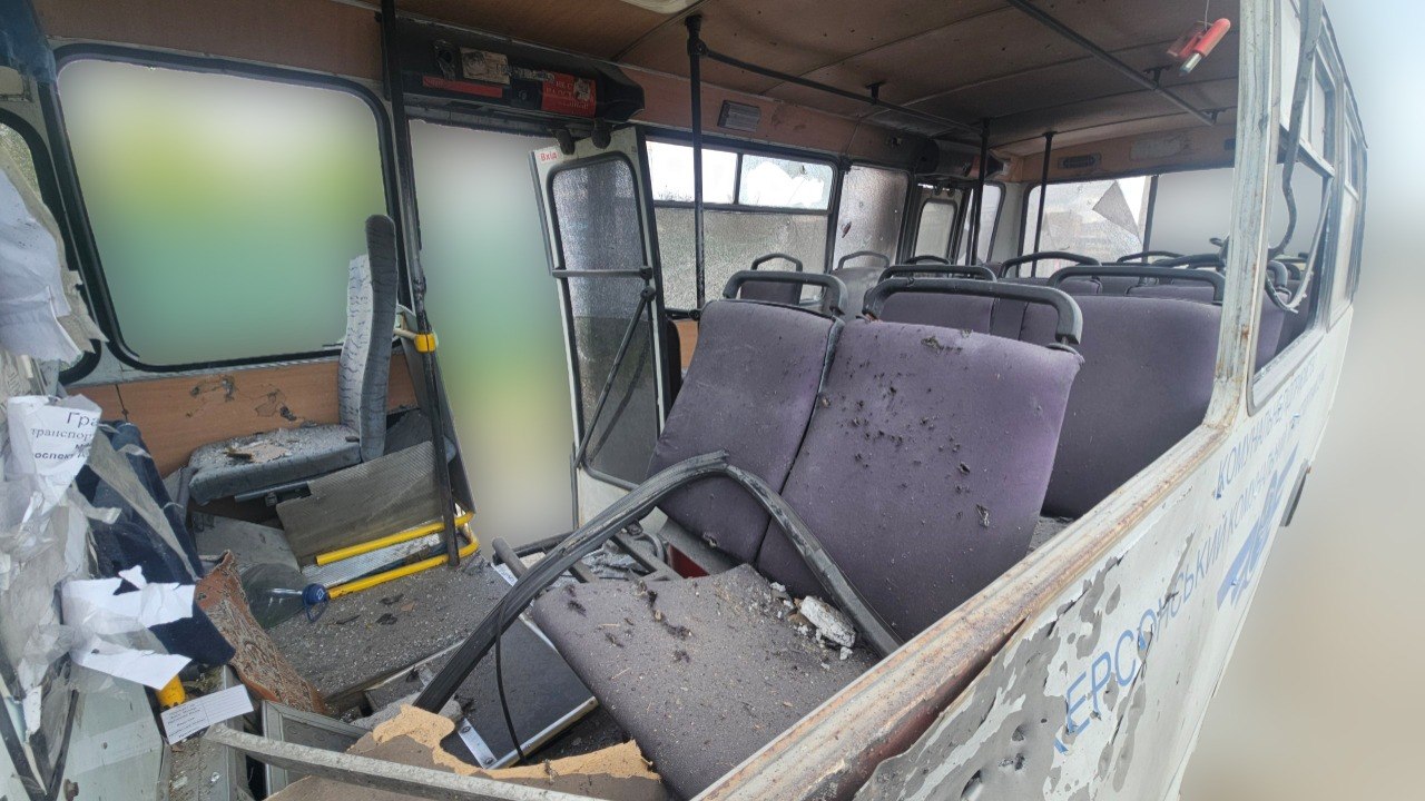 An interior view of the municipal bus shows the extent of the damage from shrapnel and glass. The purple fabric of the passenger seats is riddled with small holes, and the floor is covered in glass shards and debris. A mangled metal window frame lies across a seat in the foreground, and the exterior side of the bus is visible through the missing windows, showing heavy shrapnel pockmarks.