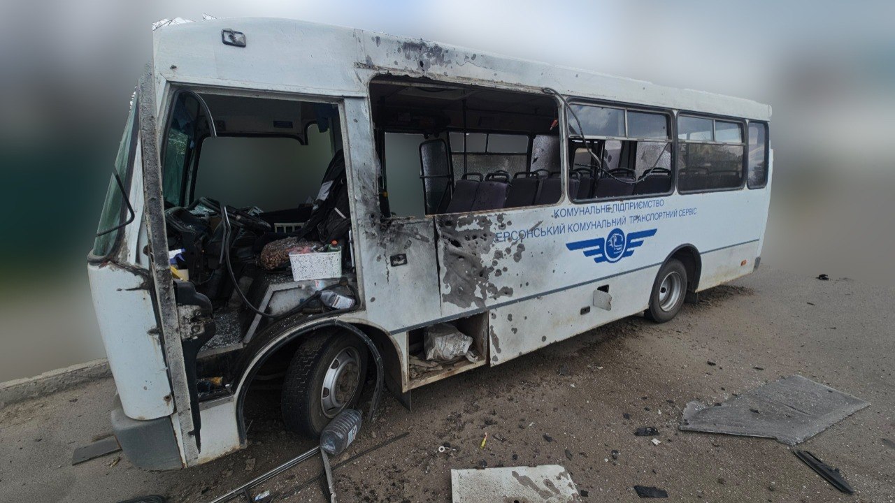 A white municipal bus from "Kherson Communal Transport Service" is shown with severe damage after an attack. The driver’s side door and several windows are missing, and the metal side of the vehicle is peppered with shrapnel holes and burn marks. Debris is scattered on the ground around the bus, and the interior seats are visible through the shattered frame.