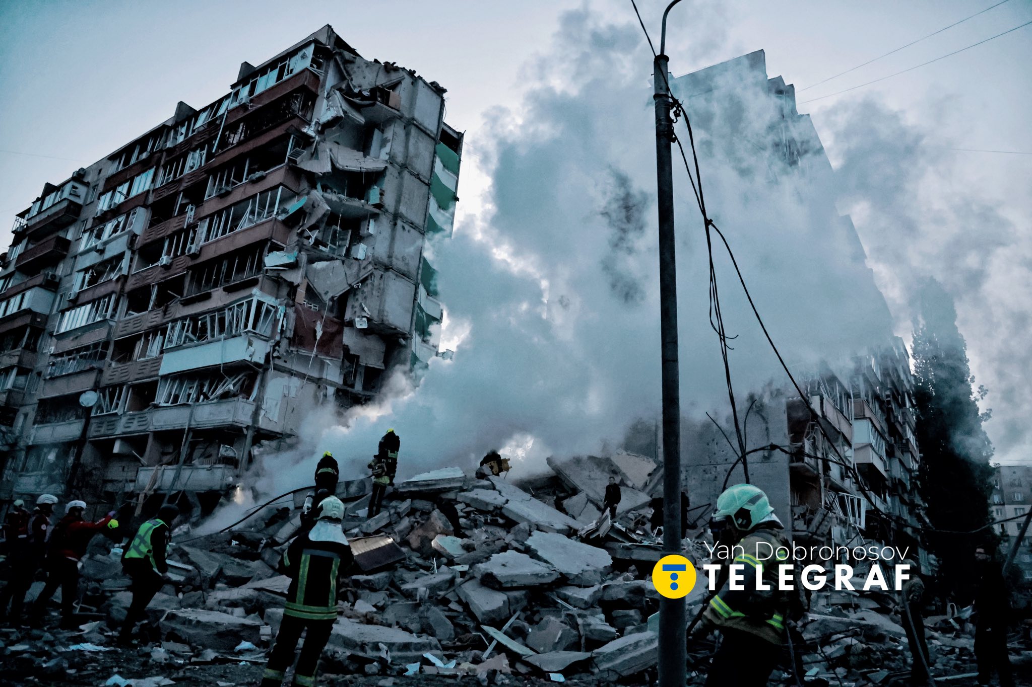A wide shot of a catastrophic scene in a residential area. A large section of a multi-story apartment building has completely collapsed, leaving a jagged vertical gap in the structure and a massive mountain of concrete rubble and twisted metal. Thick white smoke or dust billows from the ruins, partially obscuring a neighboring building. In the foreground, several first responders in high-visibility gear and helmets are working among the debris under a dim, overcast sky. A watermark for "Yan Dobronosov TELEGRAF" is in the bottom-right.