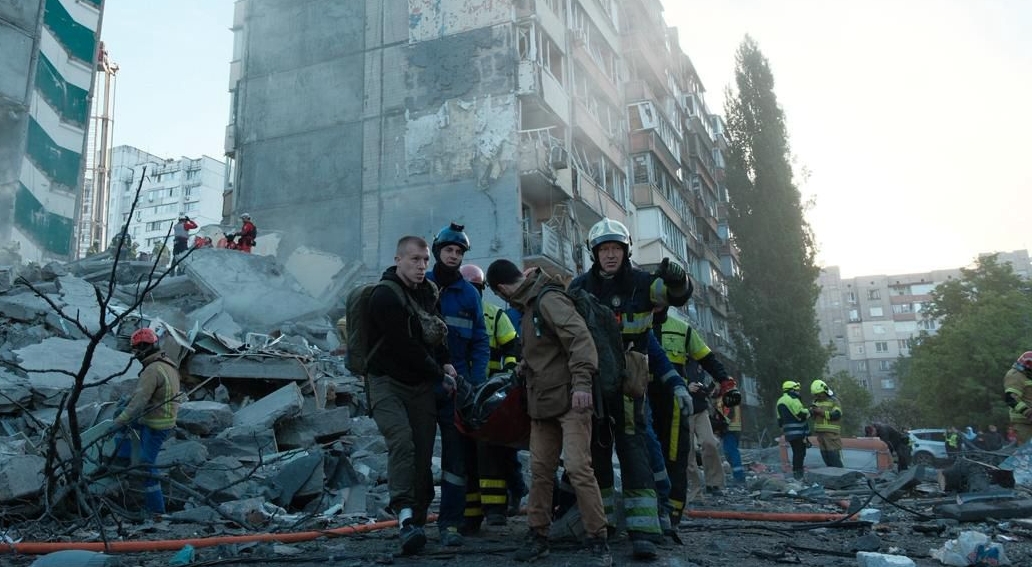 A wide shot of the aftermath of a missile strike on a multi-story residential building in Kyiv. A massive section of the apartment block has collapsed into a mountain of concrete slabs and twisted metal. In the foreground, a group of rescuers and men in civilian clothes are solemnly carrying a person on a stretcher away from the debris. Several other rescue workers in helmets and high-visibility gear are visible in the background, working among the ruins under a hazy, bright sky. The logo of the State Emergency Service of Ukraine (DSNS) in Kyiv is in the bottom-left corner.