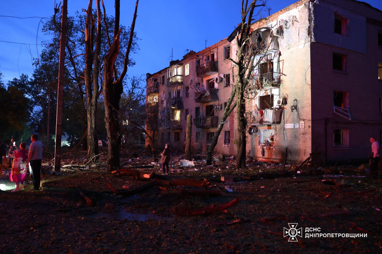 A four-story apartment building shows significant blast damage in the evening light, with blown-out windows, ruined balconies, and debris scattered across the ground. Several civilians stand nearby assessing the destruction amidst damaged trees, and a State Emergency Service logo for the Dnipropetrovsk region is visible in the bottom-right corner.