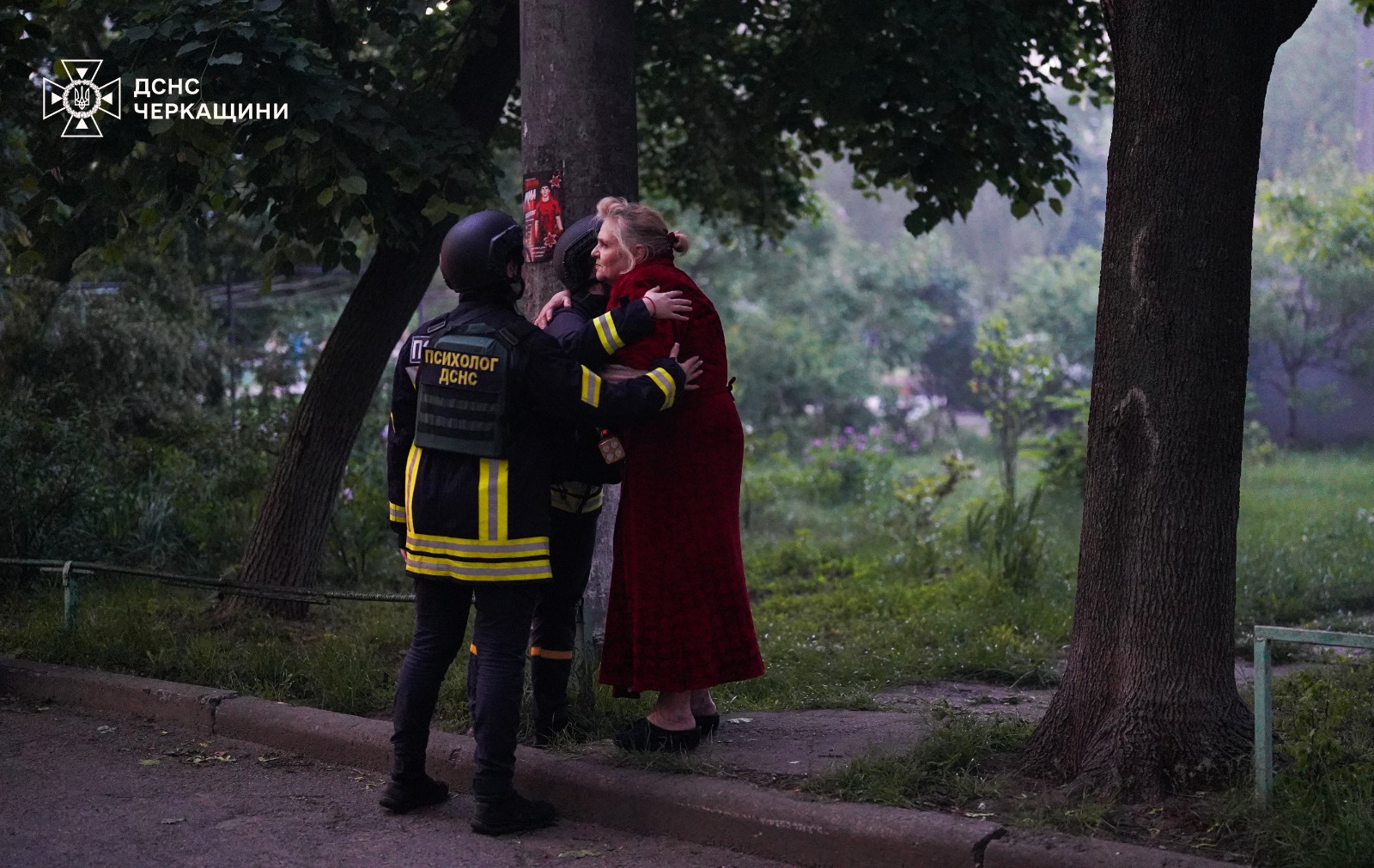 Two emergency responders in dark uniforms with yellow reflective stripes and black helmets comfort an older woman wearing a long red coat outdoors next to a tree. One of the responders has a vest patch reading "ПСИХОЛОГ ДСНС" (DSNS Psychologist), and the logo for the State Emergency Service of the Cherkasy Region is visible in the top-left corner.