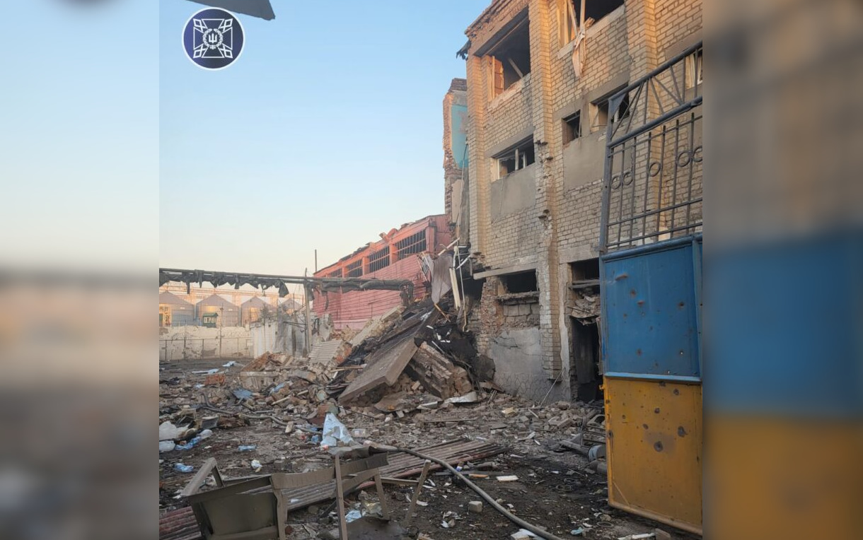 A wide shot of a multi-story brick building with severe structural damage. A large section of the exterior wall has collapsed into a massive pile of bricks and concrete debris in the foreground. The surrounding yard is littered with scattered rubble and twisted metal. In the background, a long red building and grain silos are visible under a clear sky. A blue and yellow gate stands to the right, and an official emblem is visible in the top-left corner.