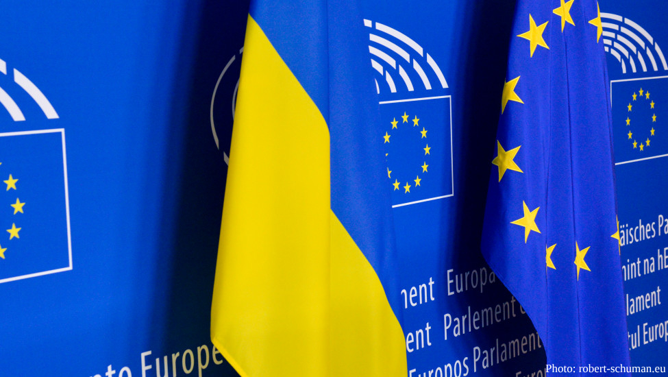 A close-up shot features the national flag of Ukraine—with its horizontal blue and yellow bands—standing alongside the blue flag of the European Union, which displays its circle of twelve gold stars. The flags are positioned against a blue backdrop that repeats the official logo and name of the European Parliament in multiple languages.