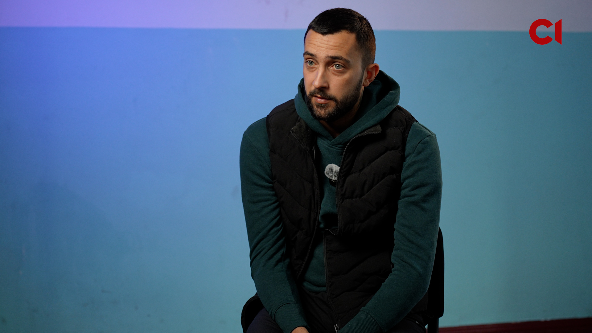 A medium shot of a man with short dark hair and a beard, looking slightly to the side with a serious expression. He is wearing a dark green hoodie under a black puffer vest with a small lapel microphone attached. The background is a plain wall with a blue and purple gradient. A red "CI" logo is in the top right corner.