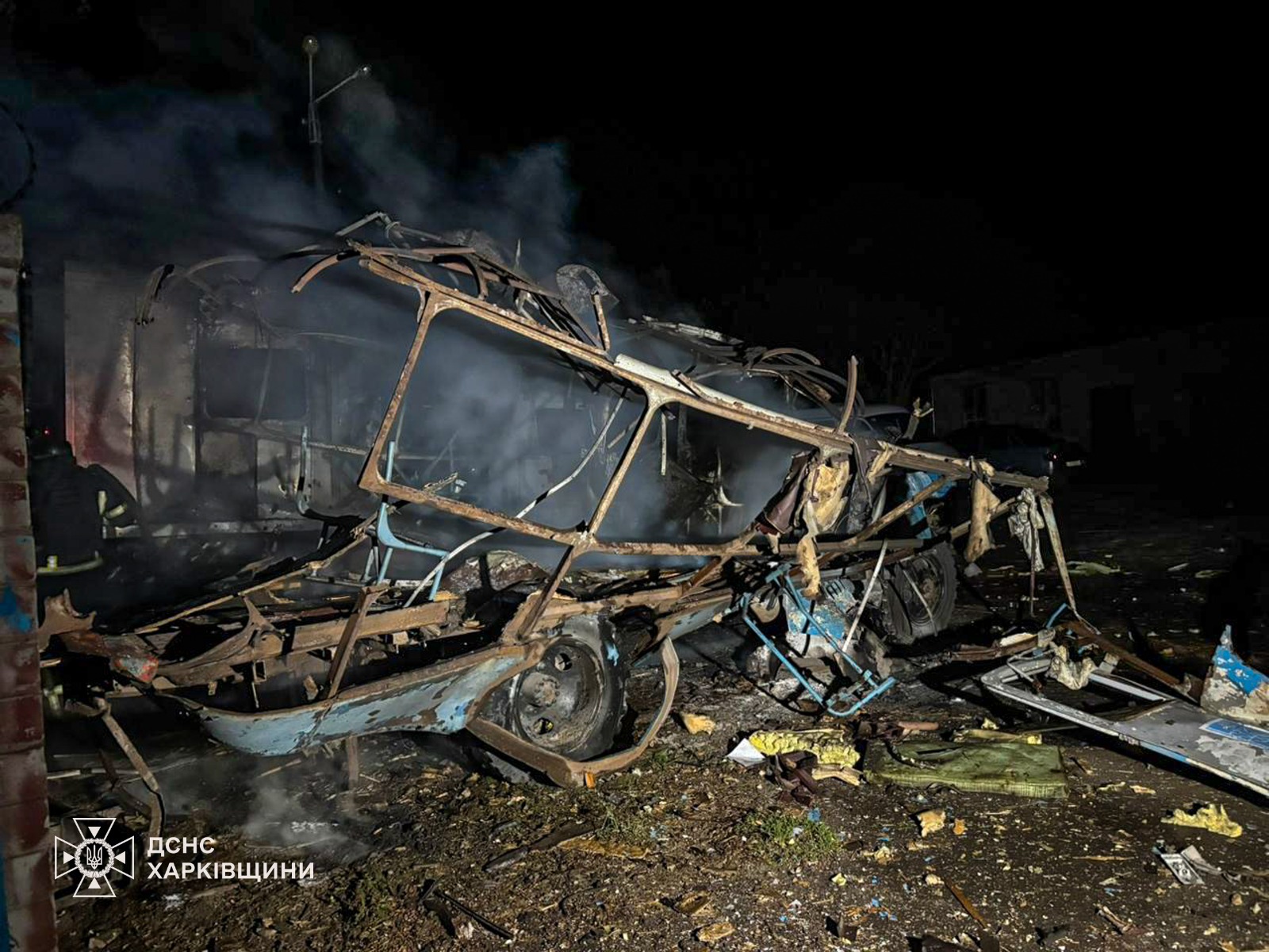 A nighttime photo shows the skeletal, burnt-out remains of a vehicle, likely a bus or large van, following an attack. Only the charred metal frame and wheels remain, with smoke still rising from the wreckage. Debris and twisted metal parts are scattered across the dark ground. In the bottom-left corner, the logo for the State Emergency Service of Ukraine (DSNS) in the Kharkiv region is visible.