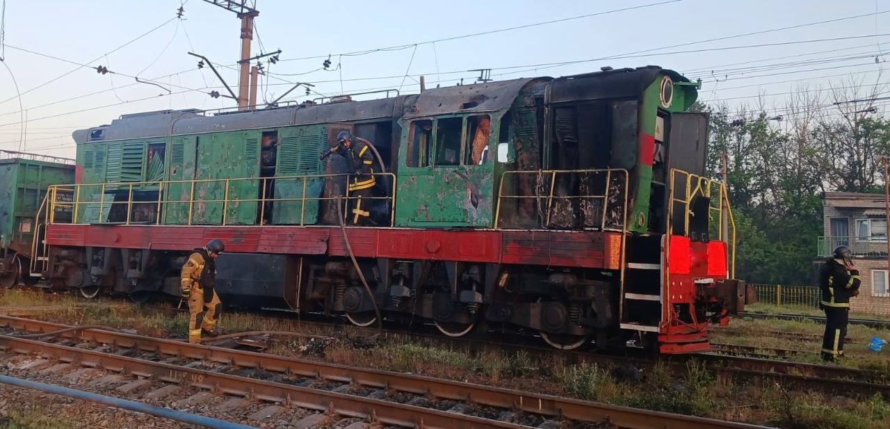 A green and red locomotive on a railway track shows significant damage, with scorched metal and shattered windows on its cab. Firefighters in protective gear are inspecting and working on the train; one is on the side walkway holding a hose, while others stand nearby on the tracks. Overhead electrical lines and a clear sky are visible in the background.
