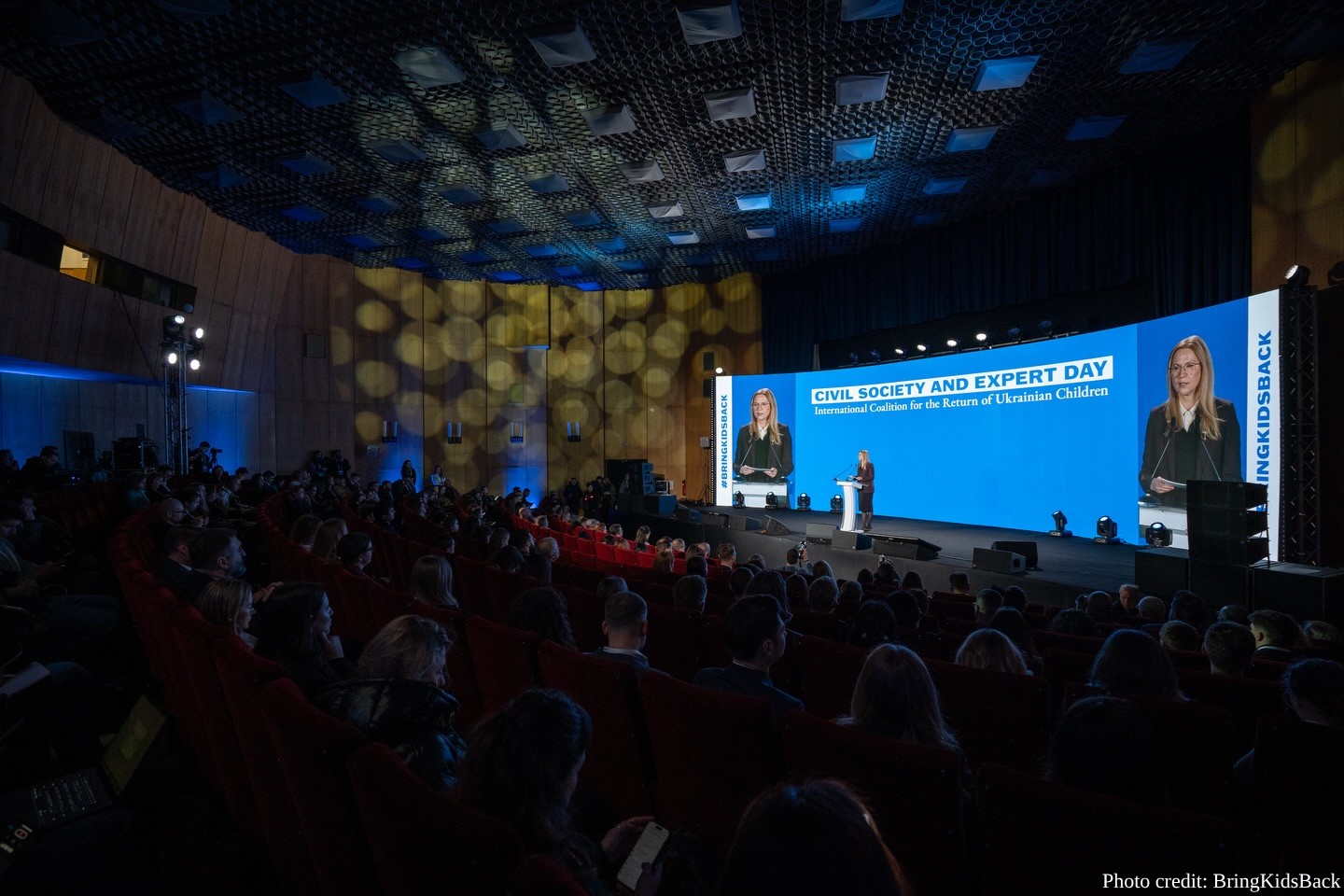A wide-angle shot of a large, dark auditorium filled with an audience seated in red chairs. On the brightly lit stage, a woman (Olena Kondratiuk) stands at a central podium speaking. A massive blue digital screen behind her displays the text "CIVIL SOCIETY AND EXPERT DAY - International Coalition for the Return of Ukrainian Children" and features live video close-ups of her on either side. The ceiling has a distinct textured, grid-like pattern, and a photo credit to "BringKidsBack" is in the bottom-right corner.